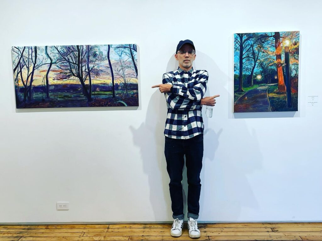 Man in checkered shirt and cap standing between two vibrant landscape paintings in an art gallery.