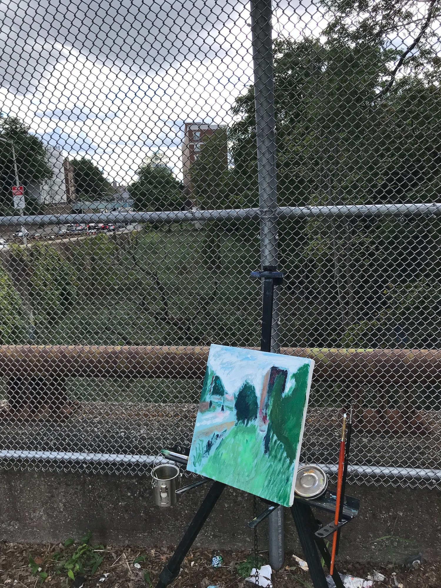 An unfinished canvas on an easel in front of a chain link fence overlooking the Tibbetts Brook area in the Bronx.