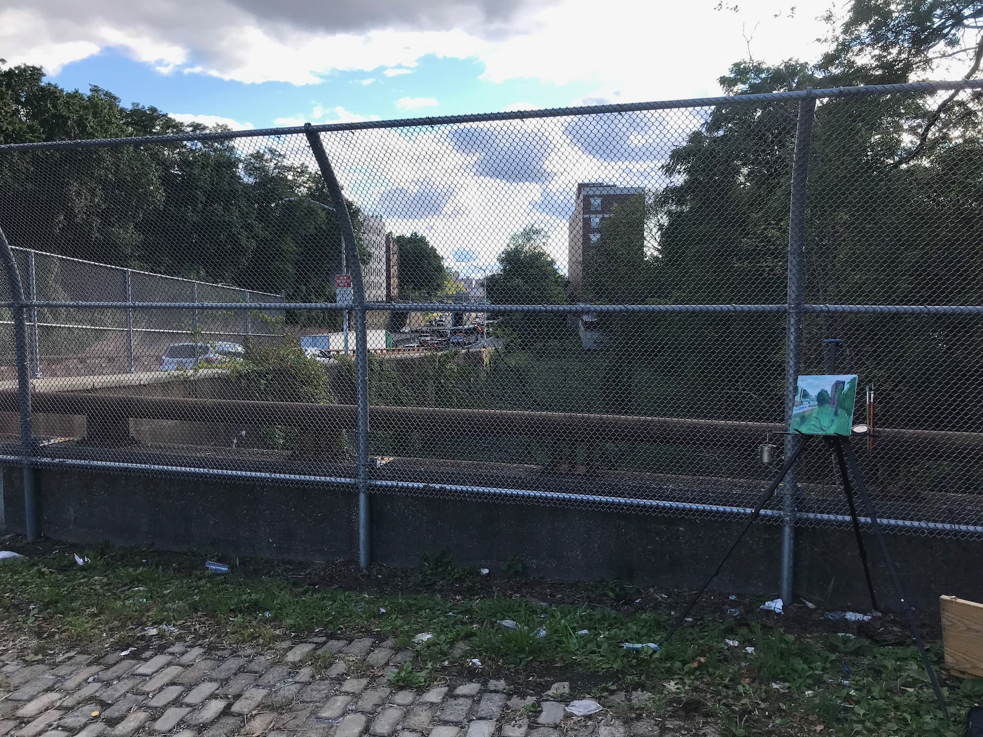 A plein air painting setup with canvas, easel, and paintbrushes against a backdrop of a chain link fence and urban greenery.
